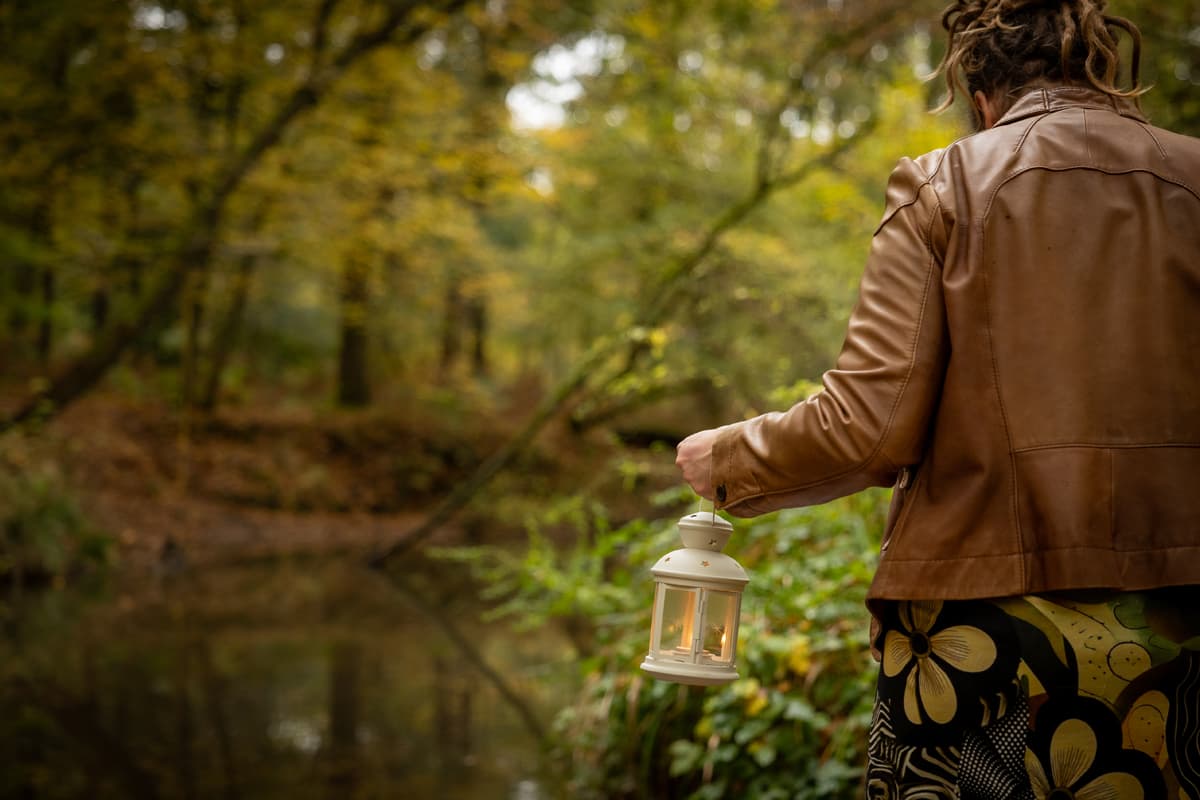 Inge staat met haar rug naar ons toe in het herfstbos en houdt een lantaarn met een brandend kaarsje bij het water